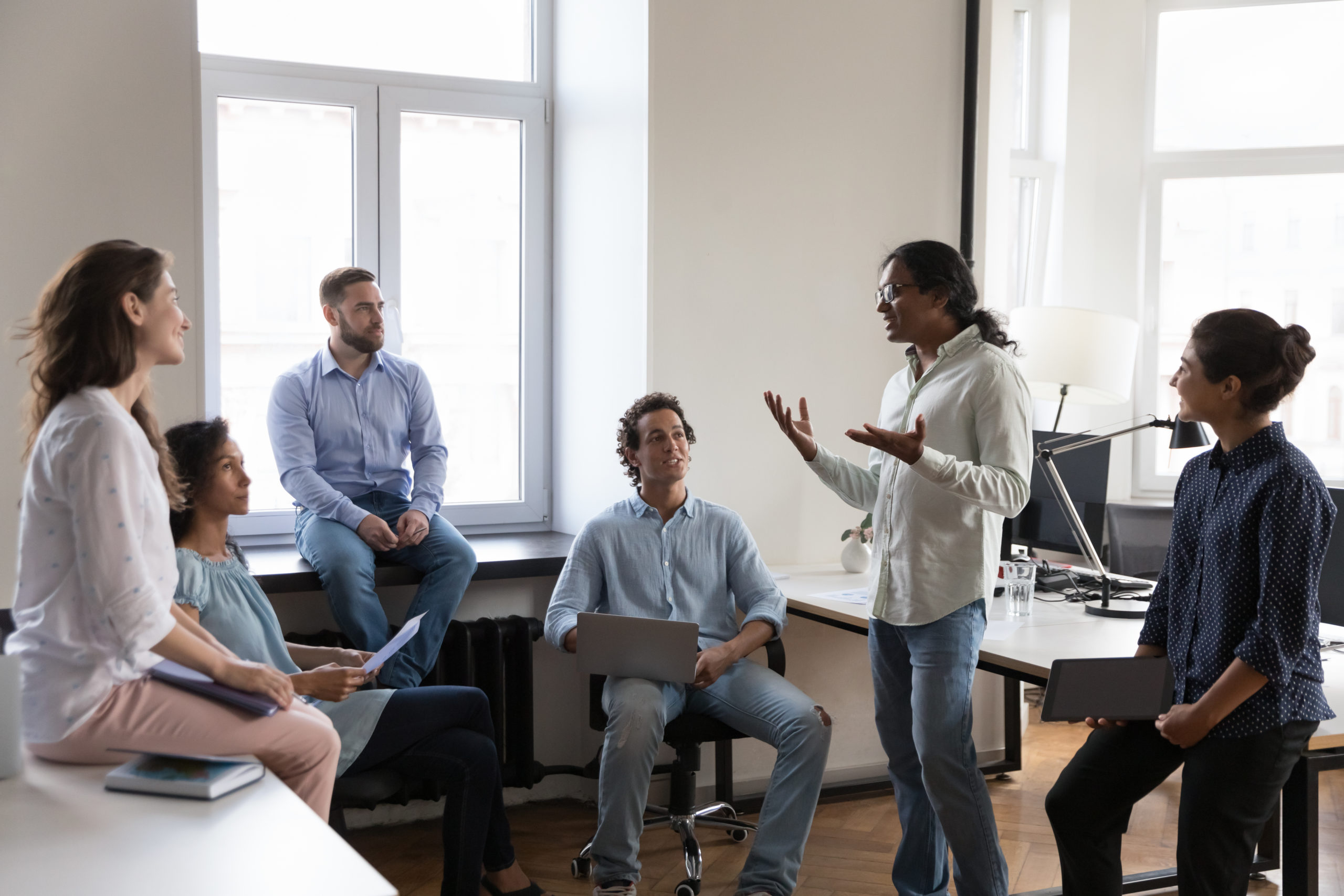 Indian businessman, coach or trainer makes speech during group briefing in co-working office with staff members, multi ethnic subordinates. Business meeting, strategizing, team-building event concept