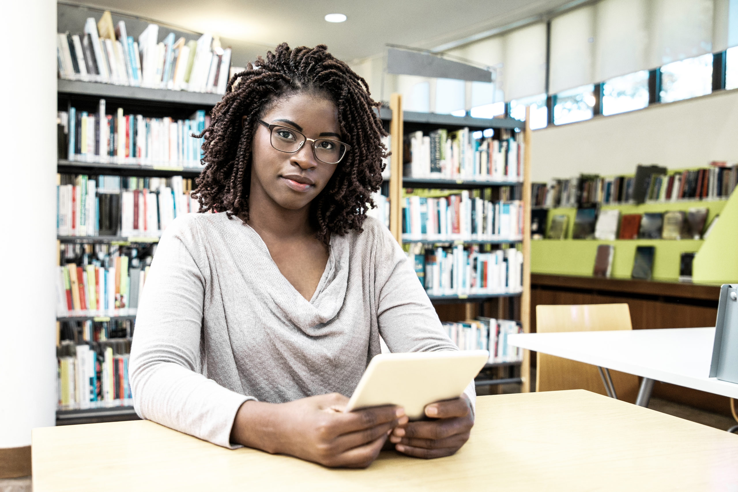 Positive female student using wireless internet connection in library. Young black woman in casual sitting at desk, using tablet and smiling at camera. Bookshelves in background. Library concept