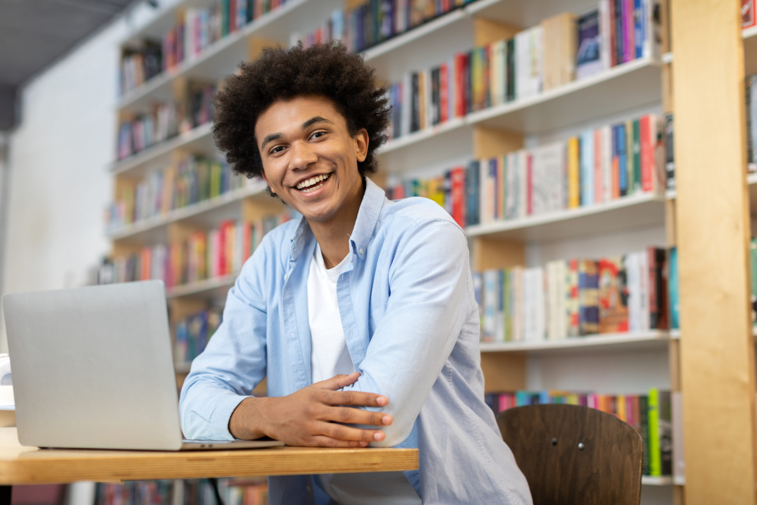 Happy black male student sitting at desk in library, in front of laptop, smiling at the camera, exuding positivity and joy in the midst of academic endeavors